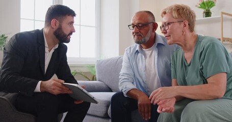 Reak estate agent consults a senior couple at home. Advisor covers finance and retirement planning in a clear consultation, using a tablet to outline options and contract points during a meeting. - Powered by Adobe
