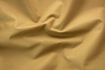 A detailed, top-down view of a golden-brown or khaki-colored fabric, showcasing its robust texture and organic folds. The material appears to be a durable synthetic textile, possibly nylon or cordura.