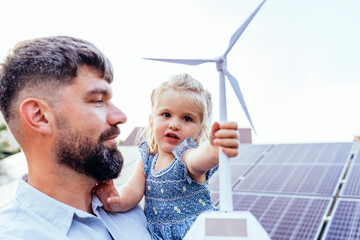 Girl holding wind turbine model while father stands near solar panels