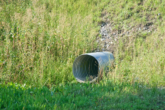 Corrugated metal drain pipe exits a grassy slope; outfall for stormwater and wastewater, guiding runoff from the hill into a ditch. Photo