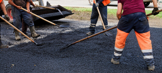 Road crew in hi-vis orange spreads and levels fresh asphalt with shovels on a city street; summer...