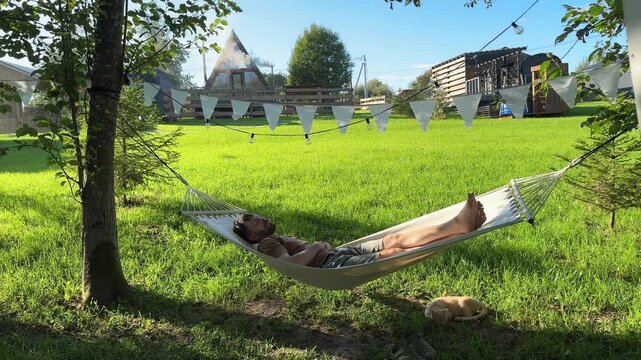 A man of Scandinavian appearance with a beard sleeps in a hammock with a garland of white flags next to a ginger cat during a light wind in the summer against the background of a country house - Powered by Adobe