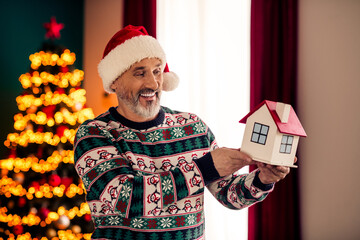 Elderly Man Holding Miniature House with Christmas Decor in Background and Joyful Holiday Atmosphere