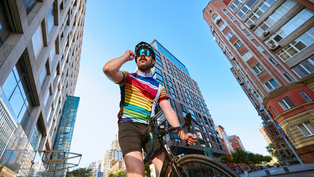 Cyclist in colorful jersey adjusting glasses with bicycle near modern buildings. Concept of urban lifestyle, sports activity, professional cycling, and emotional confidence.