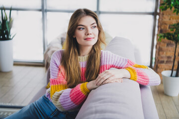 Young woman relaxing on a sofa in a cozy living room wearing casual knitwear and enjoying a...