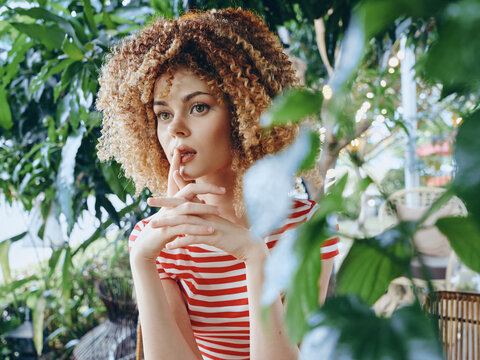 Portrait of a young woman with curly hair wearing a red and white striped top, gazing thoughtfully through lush green foliage in soft light, creating an intimate cinematic mood.