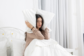 Young women covering her ears with a pillow while sitting in bed, expressing frustration, sleep...