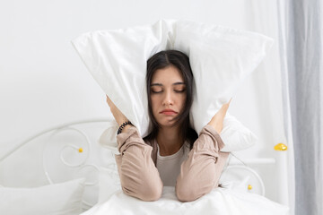 Young women covering her ears with a pillow while sitting in bed, expressing frustration, sleep disturbance, or noise problems in the bedroom, highlighting insomnia and morning irritation issues.