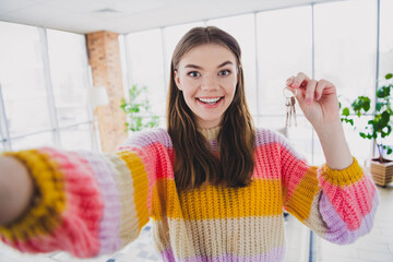 Charming young woman joyfully holding keys in a cozy modern apartment while enjoying a relaxing...