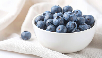 Close-up shot of a white bowl filled with fresh, ripe blueberries on a white cloth.