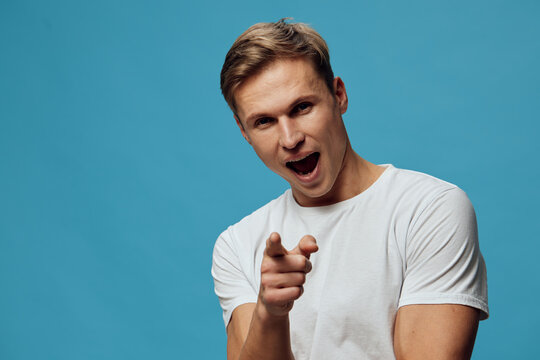 Excited young caucasian man in white t-shirt pointing finger yelling over blue background studio portrait expression concept