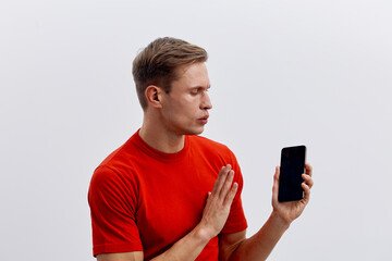 Young man with short brown hair wearing red shirt holding smartphone and making a stop gesture with his hand isolated on plain white background studio portrait. People lifestyle concept