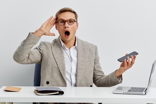 surprised man holding smartphone and laptop sitting at white desk wearing glasses gray blazer casual style isolated on light background remote work concept digital nomad communication - Powered by Adobe