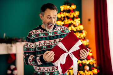 Happy senior man in festive sweater enjoying Christmas gift in a cozy holiday-decorated home with...