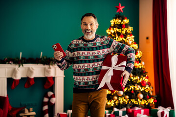 Elderly man celebrating Christmas holding a smartphone and gift box in a cozy home with festive...