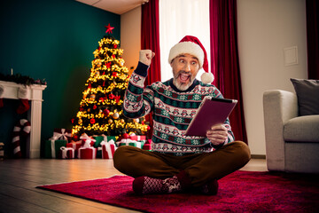 Elderly man in festive sweater celebrating Christmas using tablet in cozy holiday-decorated living...