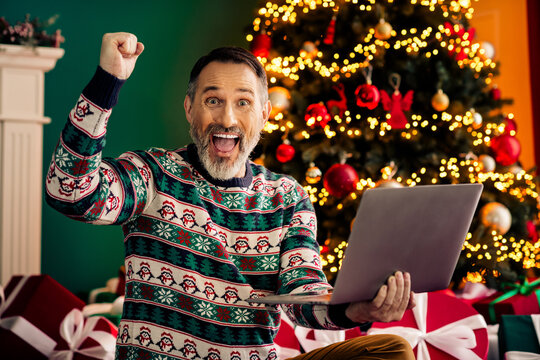 Excited senior man in festive sweater celebrating winter holiday season with laptop by illuminated Christmas tree - Powered by Adobe