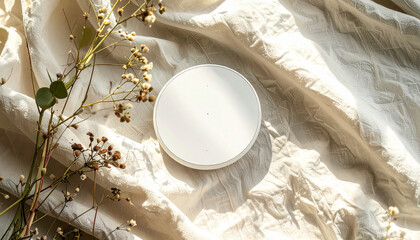 A white jar lid sits on a wrinkled white cloth, with delicate flowers and leaves nearby.