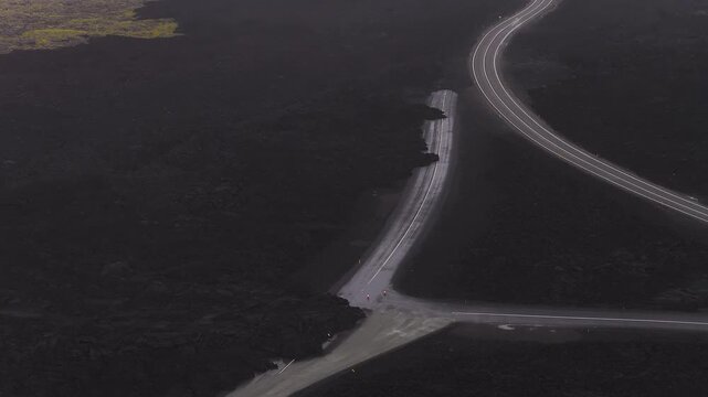 Aerial view of a winding road cutting through dark lava fields with green moss patches, showing partial destruction and gradual panning motion.