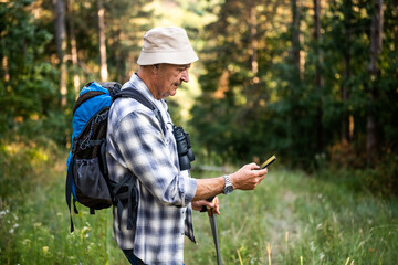 Portrait of active senior man with backpack and hiking poles using mobile phone while enjoying hiking in nature.