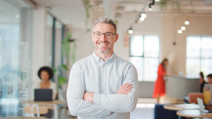 Portrait Of Smiling Mature Businessman Wearing Glasses Standing In Modern Open Plan Office
