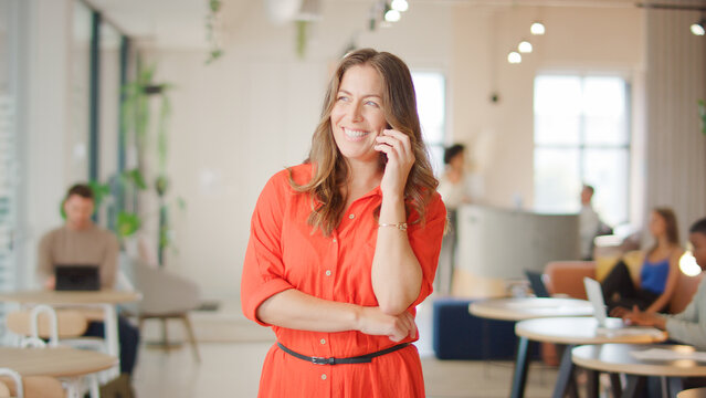 Smiling Mature Businesswoman Standing In Busy Modern Open Plan Office Talking On Mobile Phone