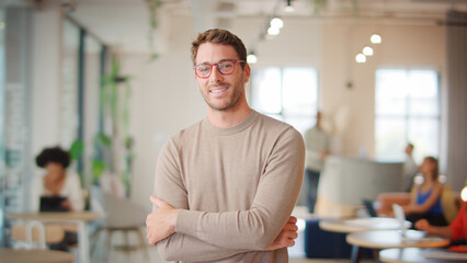 Portrait Of Smiling Businessman Wearing Glasses Standing In Busy Modern Open Plan Office