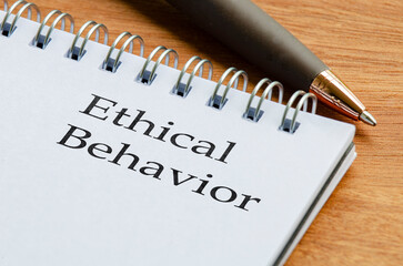 Close up notepad with the words Ethical Behaviour written on a page placed on a wooden desk with a...
