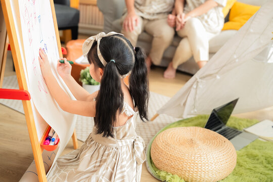 Happy Asian family spending quality time together at home. Father and daughter drawing on the floor with colored pens while mother works on a digital tablet smartphone on the sofa. Warm, modern living