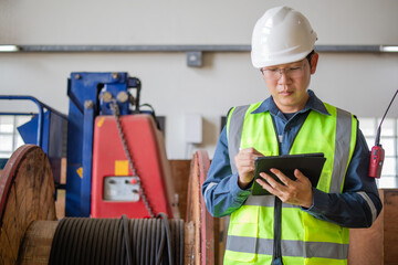 Engineer in factory conducts powercable inspection using tablet. Wearing hard hat and safety vest, he ensures safety and efficiency in industrial environment