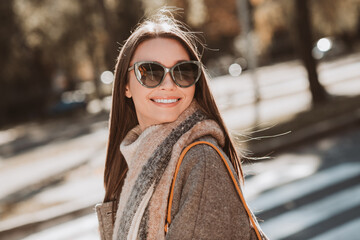 Glamorous woman in chic autumn attire smiling outdoors in a sunny urban setting with a picturesque...