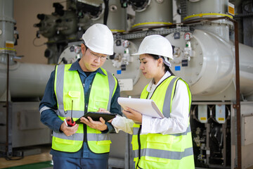 Two factory workers wearing safety helmets and reflective vests demonstrate teamwork while checking energy equipment and discussing plans to ensure efficient factory operation and safety