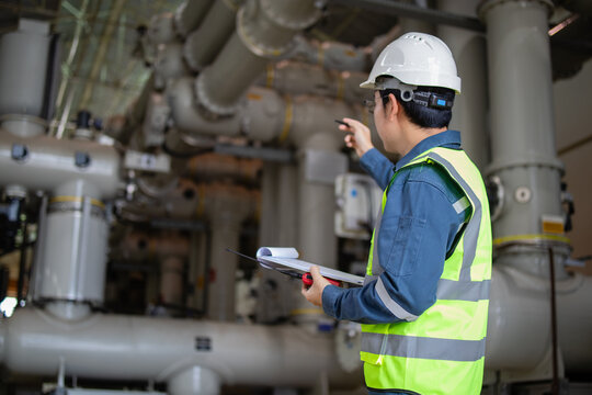 Worker in safety vest and helmet performing electrical inspection at power plant with clipboard and pen checking industrial pipes and equipment carefully - Powered by Adobe
