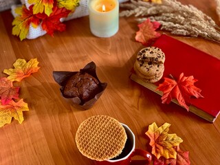 A warm and inviting table displays a muffin, cookies, and a waffle. Bright autumn leaves surround a flickering candle and a book, creating a cozy ambiance perfect for fall