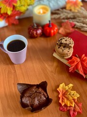 Warm coffee sits next to chocolate chip cookies and a muffin, surrounded by autumn leaves and small decorative pumpkins, creating a cozy fall atmosphere