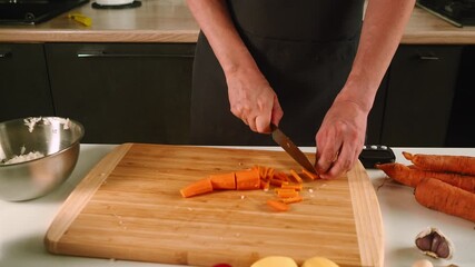 In a modern kitchen, someone skillfully chops bright orange carrots on a wooden cutting board. Fresh vegetables and a mixing bowl suggest an emphasis on healthy cooking and meal preparation