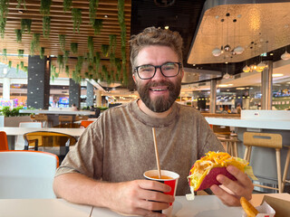 A bearded man with glasses smiles while holding a taco and drink inside a restaurant. Dining...