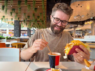 A man with glasses enjoys a taco and drink while sitting inside a food court restaurant. Casual...