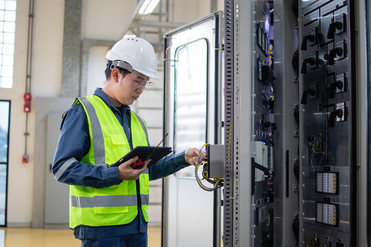 Electrical engineer wearing safety helmet and reflective vest performing inspection on substation control panel using digital tablet in industrial facility