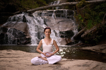 Young woman sitting in lotus pose under tropical waterfall meditating with calm strength and mindfulness fully connected to nature energy tranquility and inner balance