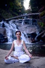 Young woman sitting in lotus pose under tropical waterfall meditating with calm strength and mindfulness fully connected to nature energy tranquility and inner balance