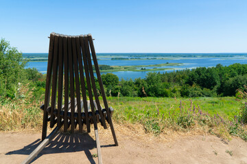 A rustic wooden folding chair on a grassy hill, offering a panoramic view of a wide river and a lush green forest under a clear blue sky