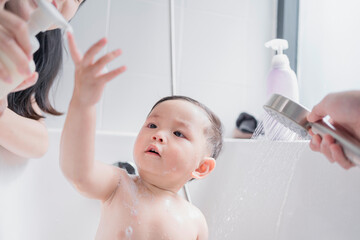 Asian parents bathing their toddler in a clean, modern bathroom. Father using handheld shower while...