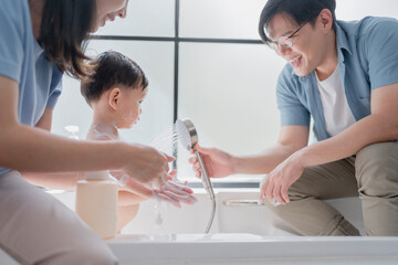 Asian parents bathing their toddler in a clean, modern bathroom. Father using handheld shower while...