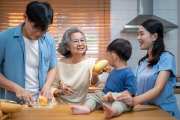 Fototapeta premium Asian family spending quality time in the kitchen — a grandmother playfully bites into a mango while surrounded by her son, daughter-in-law, and grandson during food preparation.