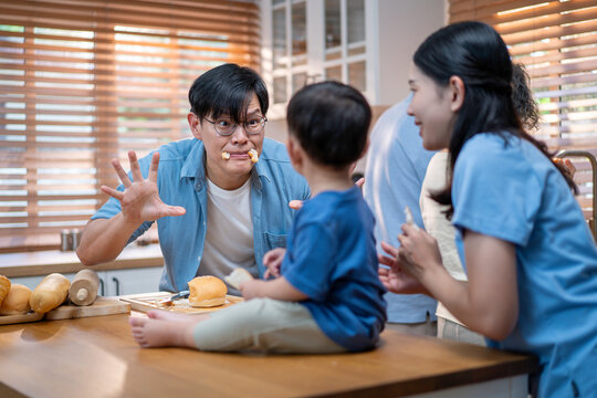 Asian family preparing food together in a cozy kitchen  a father slicing bread while the mother and child watch joyfully, surrounded by three generations. - Powered by Adobe