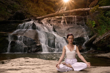 Young woman sitting in lotus pose under tropical waterfall meditating with calm strength and mindfulness fully connected to nature energy tranquility and inner balance