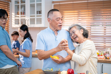 Fototapeta premium Happy Asian senior couple preparing fruit together in the kitchen while sharing laughter and love, surrounded by family in the background. A heartwarming moment of connection and healthy living.