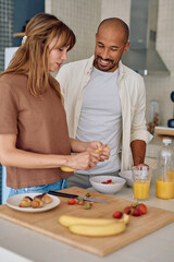Young multi-ethnic couple preparing healthy breakfast with fresh fruit and orange juice, smiling and looking at each other in modern kitchen
