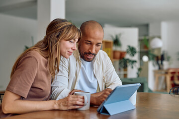 Smiling couple enjoying coffee and browsing on a digital tablet while seated at a table in their cozy living room, sharing moments together